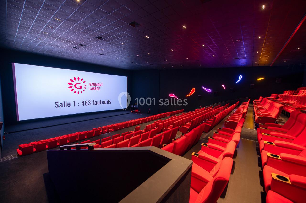 Salle de cinéma avec des sièges rouges, un grand écran affichant ’Salle 1 : 483 fauteuils’ et des lumières décoratives…