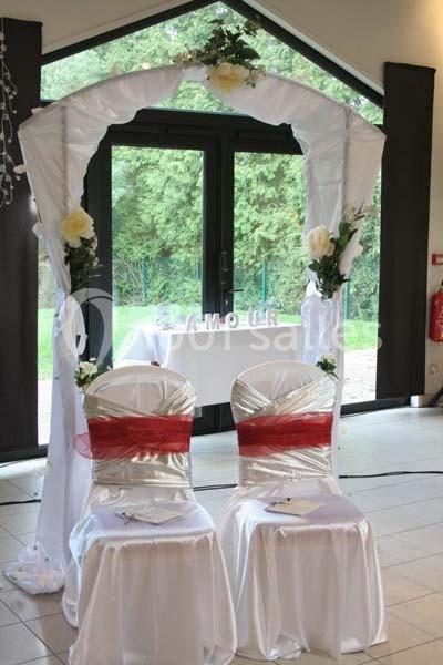 Deux chaises décorées devant une arche fleurie blanche, installées dans une salle avec vue sur un jardin.