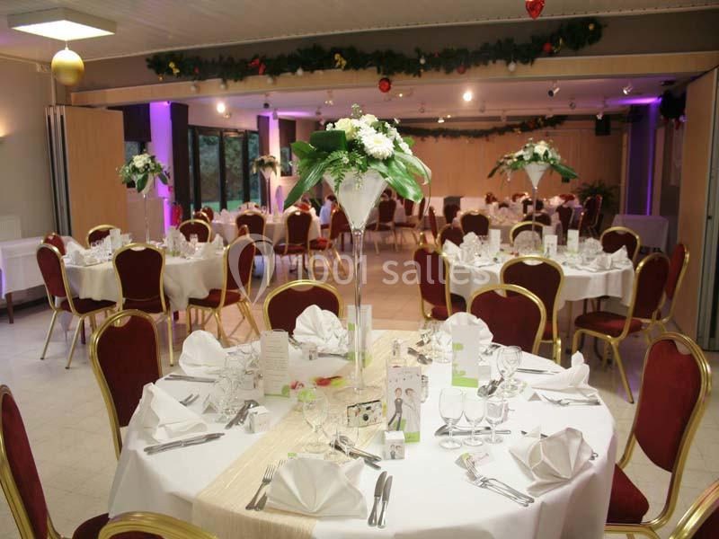 Salle de réception décorée avec des tables rondes, nappes blanches, chaises rouges et centres de table floraux.
