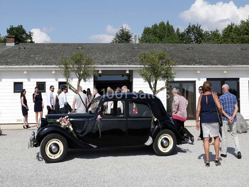 Voiture ancienne noire décorée pour un mariage, stationnée devant un bâtiment blanc, entourée de plusieurs personnes.
