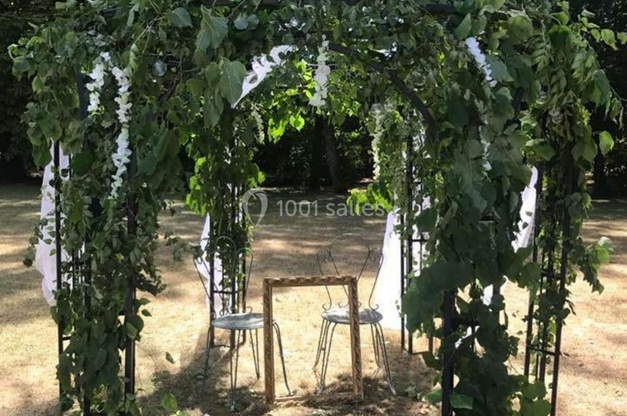 Tonnelle décorée de feuillage et de fleurs blanches, abritant deux chaises et une petite table en métal, dans un jardin.