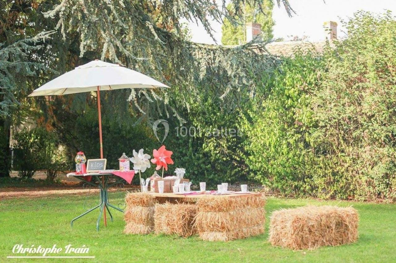 Table basse en bottes de paille avec décoration champêtre, sous un parasol blanc dans un jardin verdoyant.