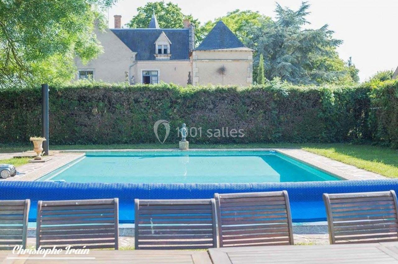 Piscine extérieure entourée de haies, avec une maison en pierre et des arbres en arrière-plan.