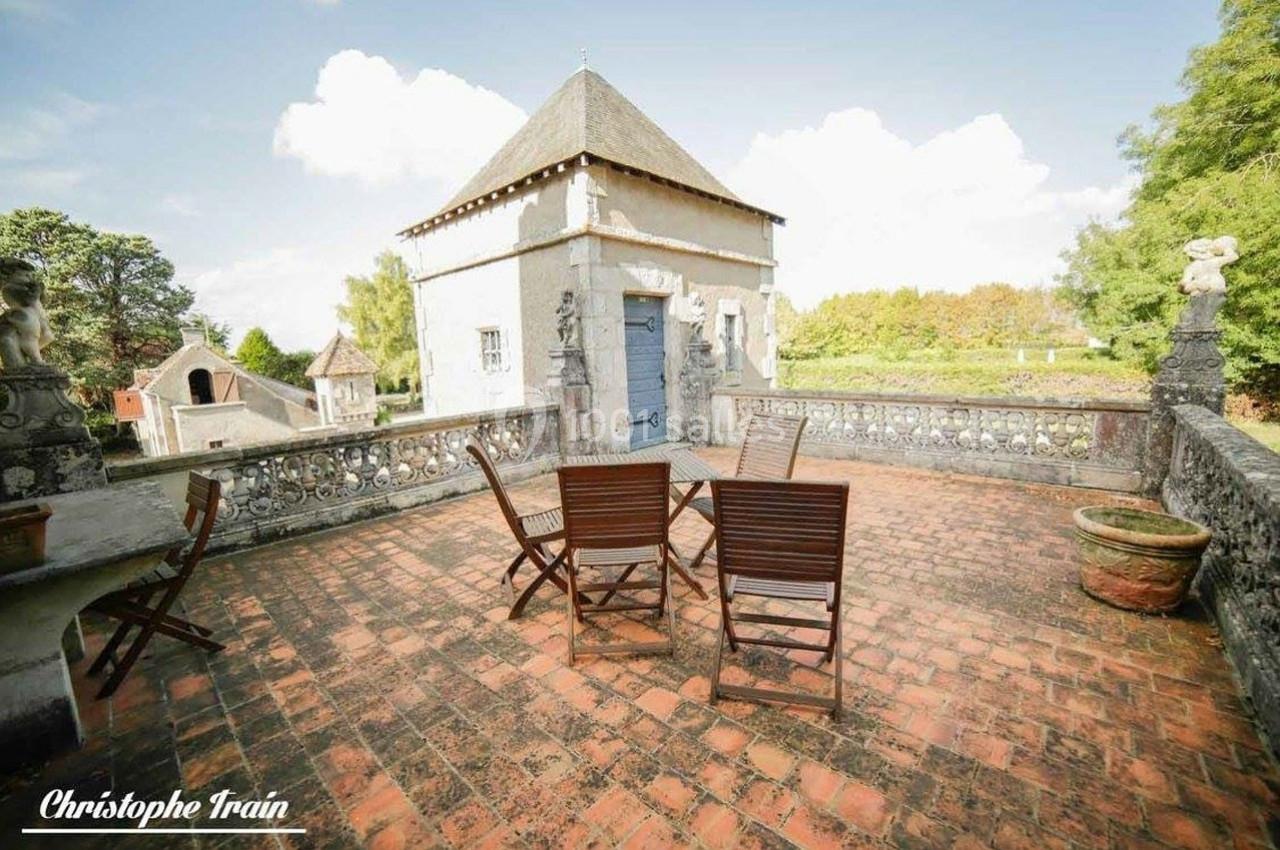 Terrasse en briques avec table et chaises en bois, entourée de balustrades et donnant sur un bâtiment ancien.