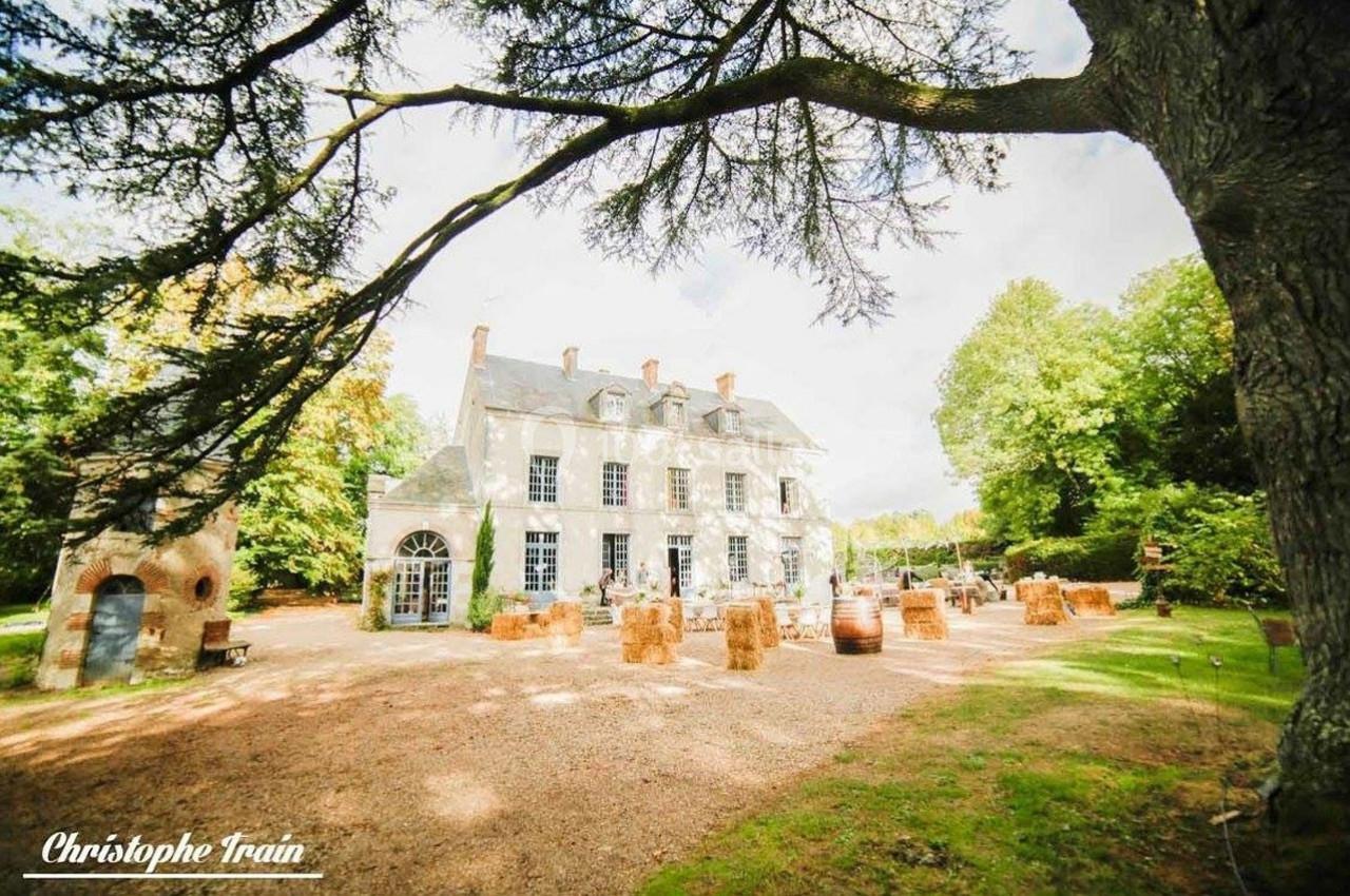 Grande maison de campagne entourée d'arbres, avec une cour aménagée de bottes de foin et de tonneaux.