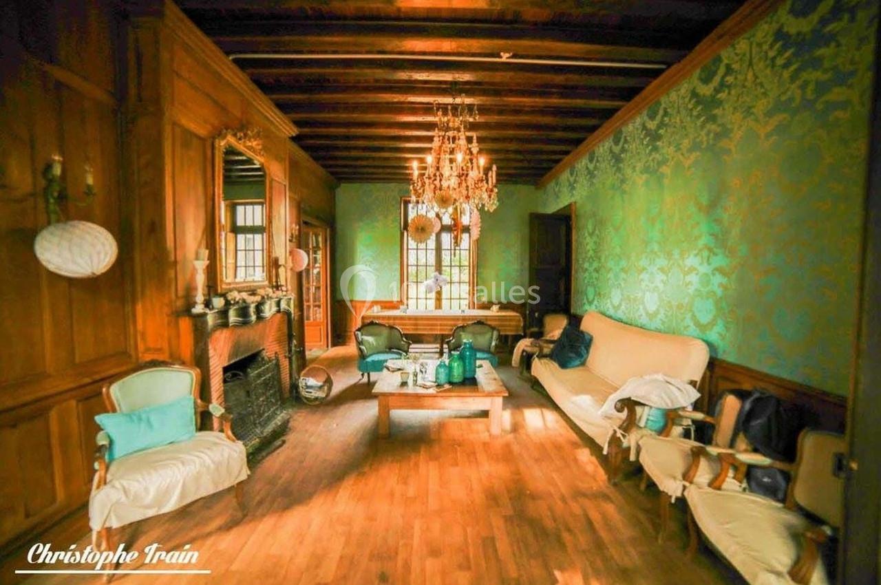 Salon avec murs en bois et papier peint vert, plafond à poutres apparentes, lustre, cheminée et mobilier ancien.