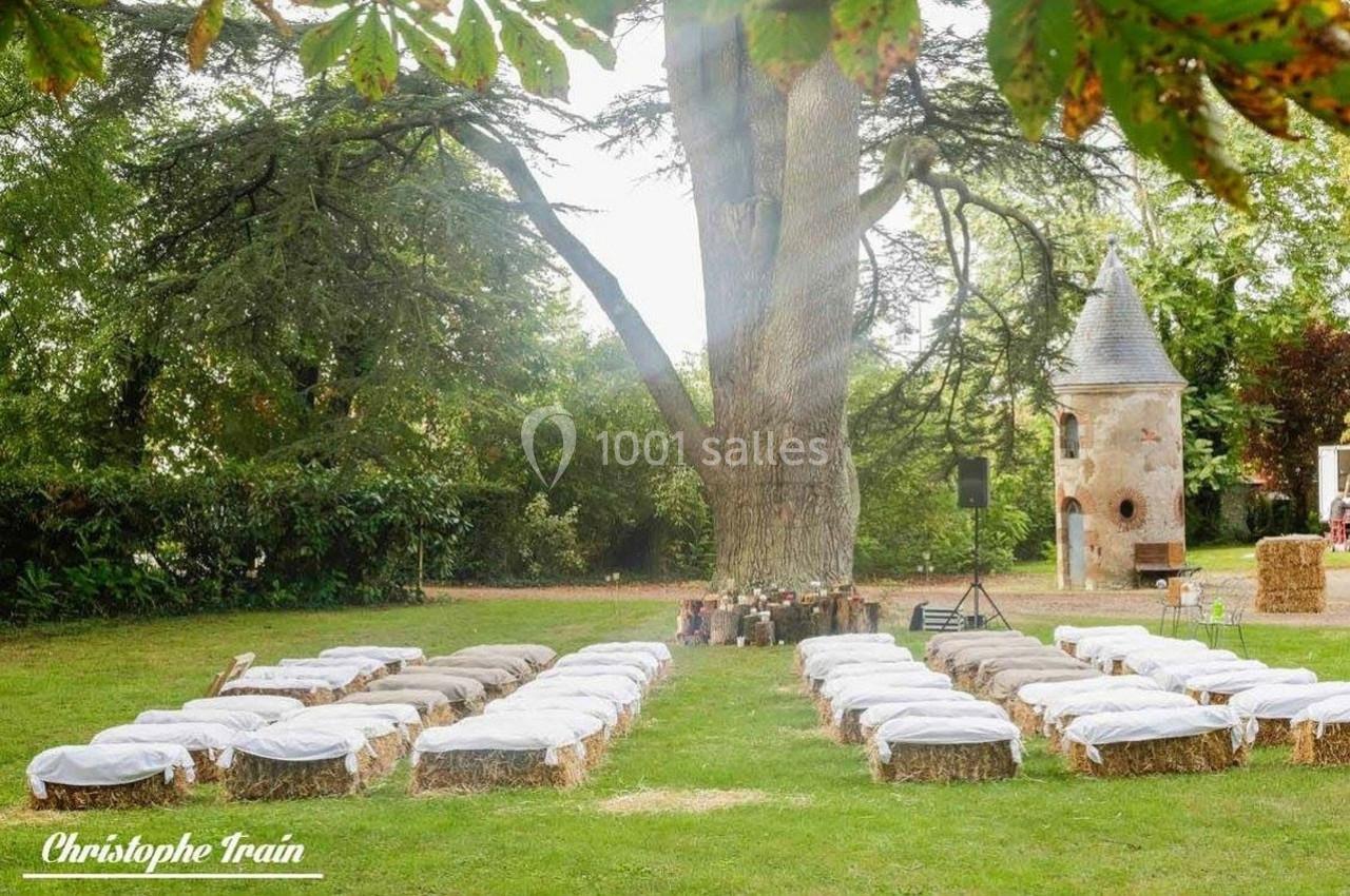 Chaises en bottes de paille recouvertes de tissu blanc disposées en extérieur sous un grand arbre, près d'une tour en pierre.