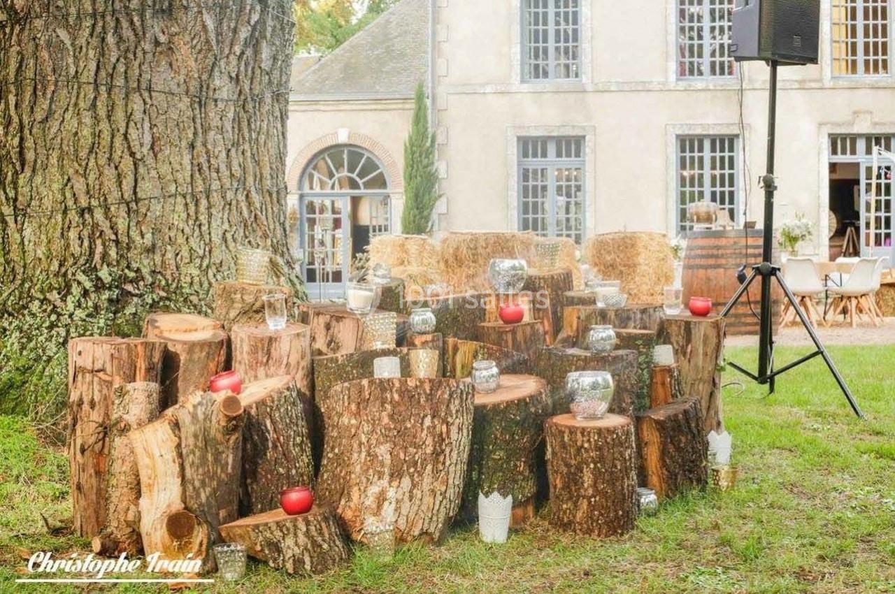 Des rondins de bois empilés décorés de bougies et photophores, disposés dans un jardin près d'un bâtiment ancien.