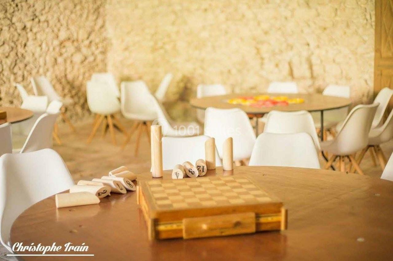 Jeu de société en bois posé sur une table ronde, entourée de chaises blanches dans une salle lumineuse en pierre.