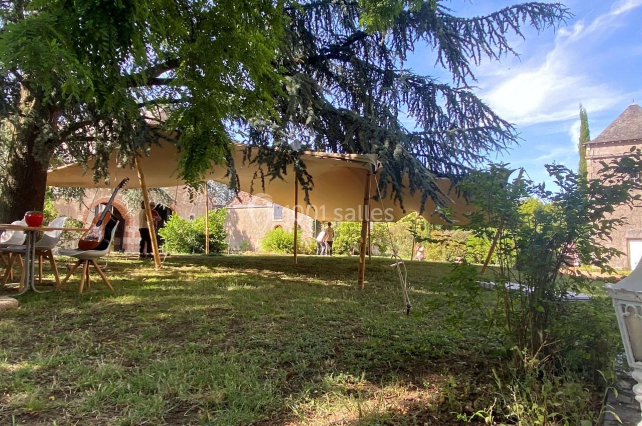 Grande tente beige installée dans un jardin ombragé, entourée de chaises et de tables sous des arbres.