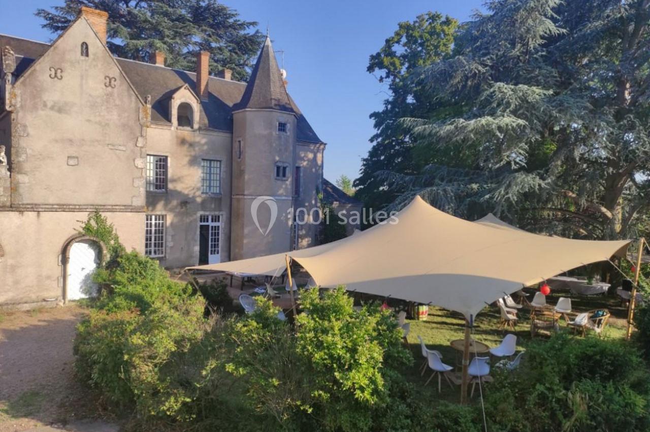 Façade d’un château ancien avec une tente beige installée dans le jardin, entourée de végétation et de mobilier extérieur.