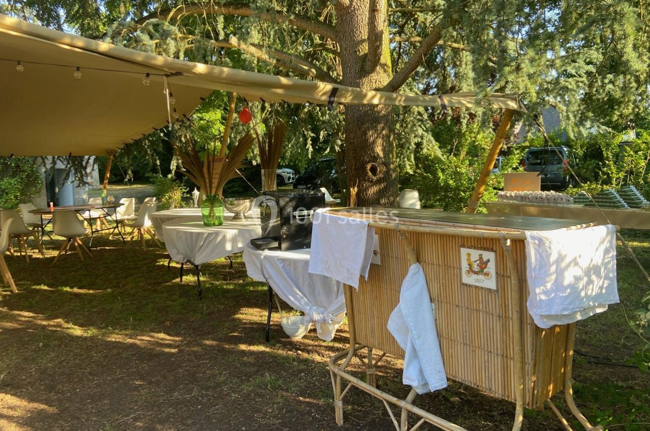 Espace extérieur ombragé avec tables, chaises et un comptoir en bambou sous un grand arbre.