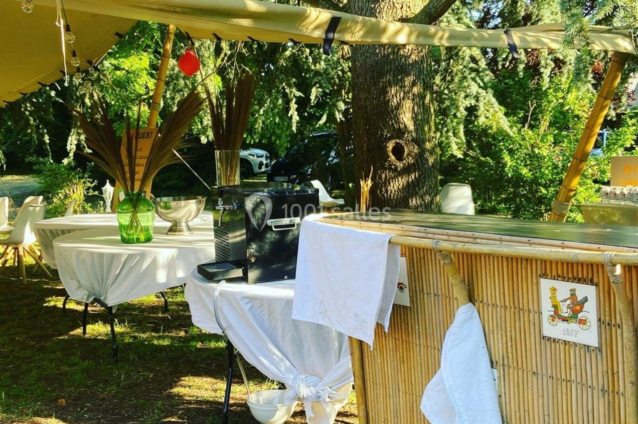 Stand extérieur avec bar en bambou, tables décorées et matériel sous une tente, entouré de végétation.