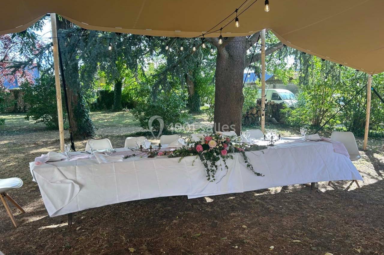 Table dressée en extérieur sous une tente, ornée d'une nappe blanche et d'une décoration florale au centre.