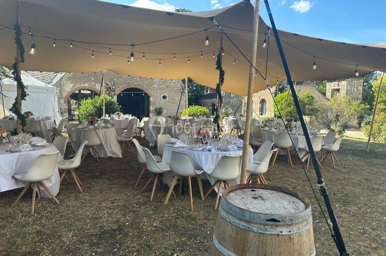 Tables dressées sous une tente beige avec guirlandes lumineuses, dans un jardin près de bâtiments en pierre.