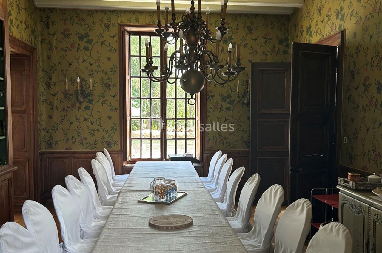 Salle à manger avec une longue table couverte d'une nappe blanche, entourée de chaises et éclairée par un lustre ancien.