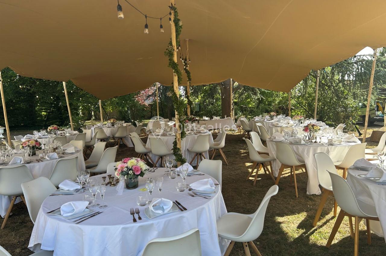 Tables rondes dressées avec nappes blanches, chaises modernes et décor floral sous une tente en extérieur.