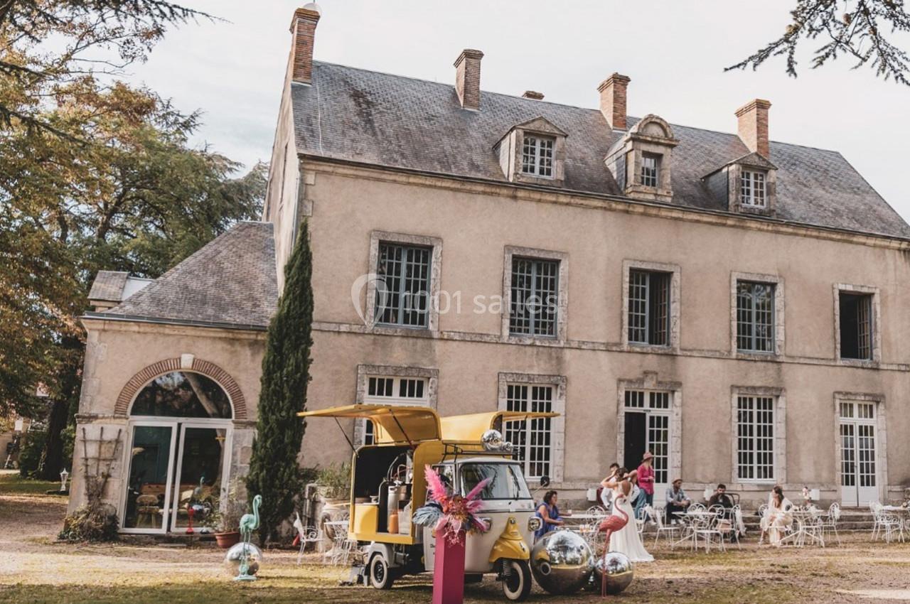 Façade d'un manoir ancien avec des tables en extérieur, un triporteur jaune et des décorations colorées au premier plan.