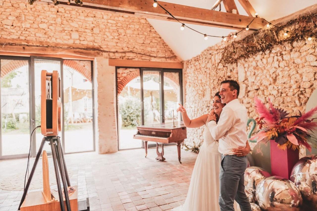 Un couple souriant utilise une borne photo dans une salle en pierre décorée de guirlandes lumineuses et de fleurs colorées.