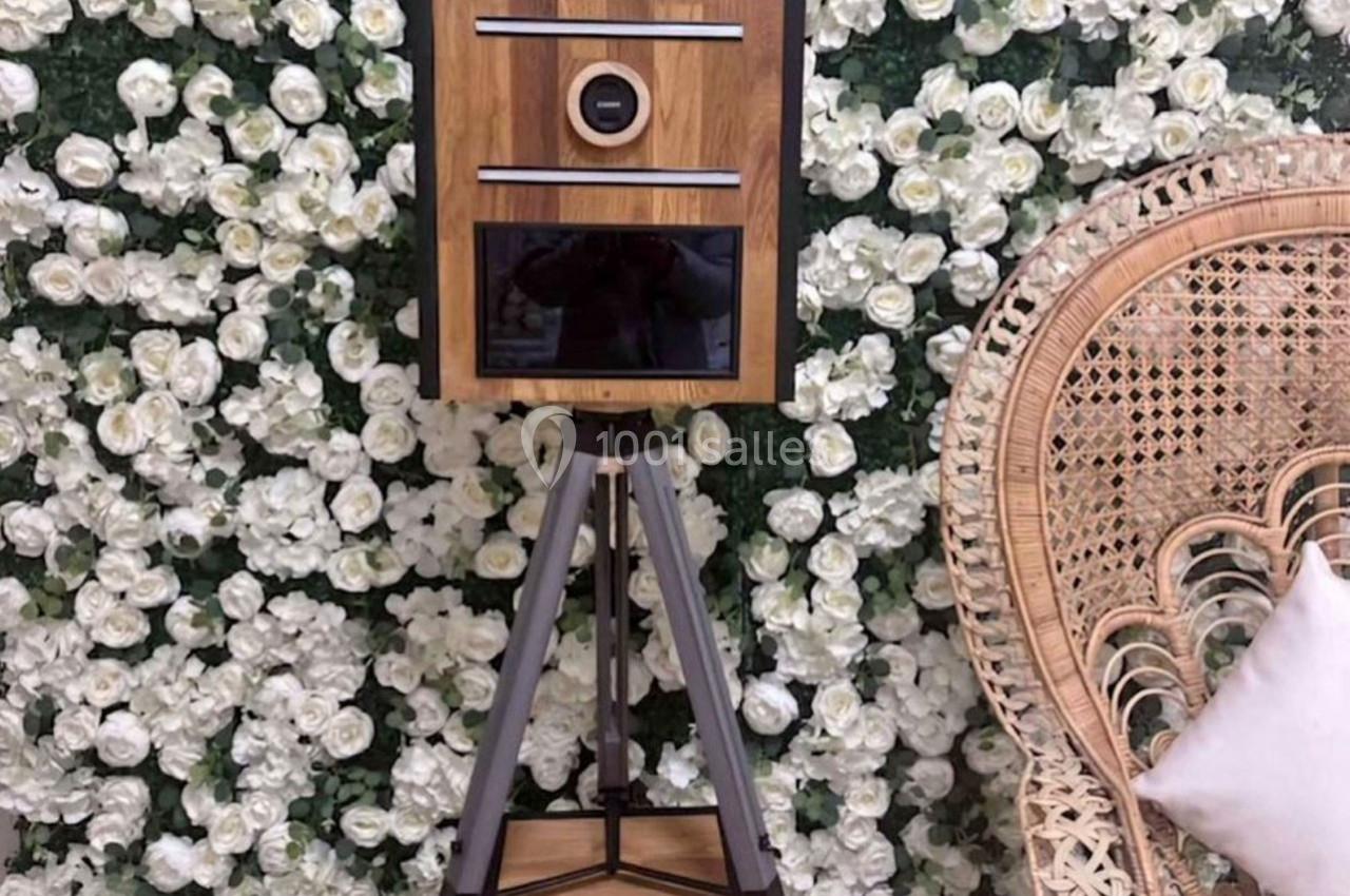 Borne photo en bois sur trépied devant un mur de fleurs blanches et un fauteuil en rotin.