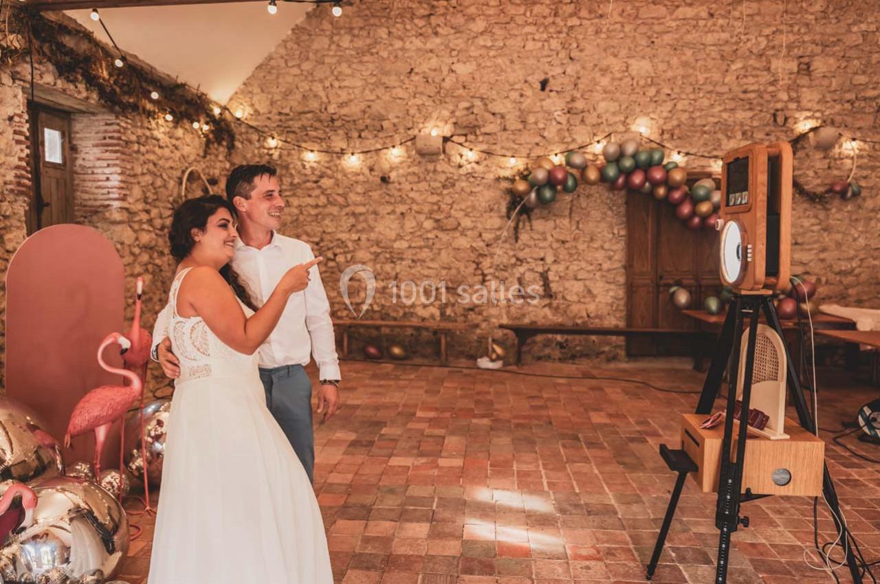 Un couple souriant pose devant une borne photo dans une salle en pierre décorée de guirlandes lumineuses et ballons.