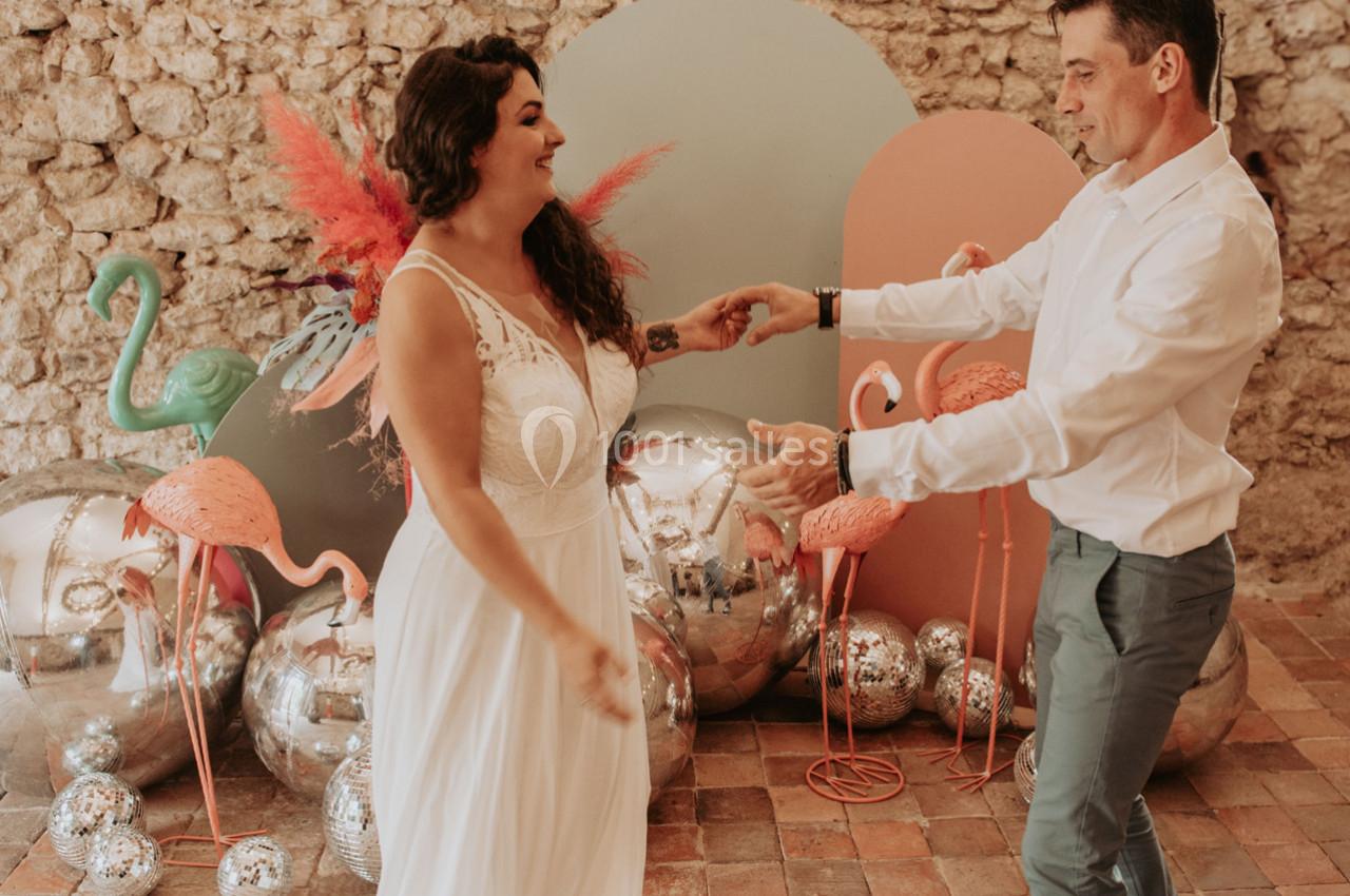 Un couple danse joyeusement dans un décor coloré avec des flamants roses, des ballons et un mur en pierre.