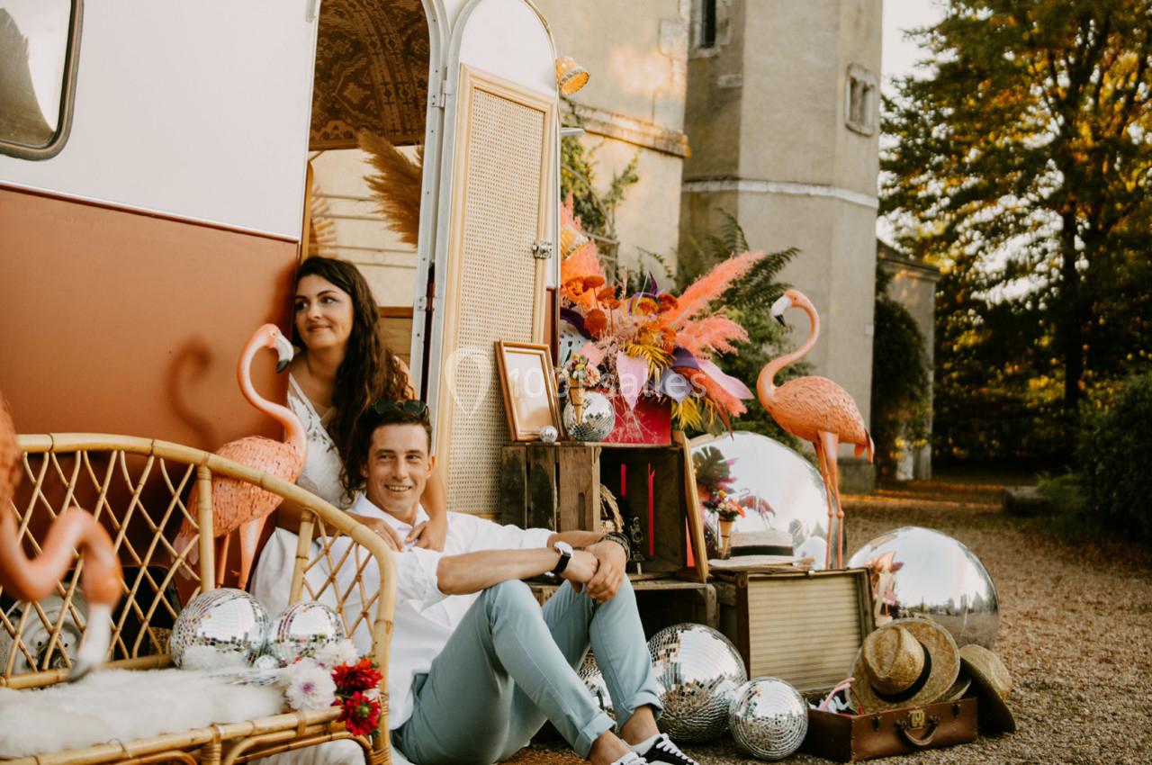 Un couple posé devant un van rétro décoré de flamants roses, miroirs et fleurs, près d'un bâtiment ancien.