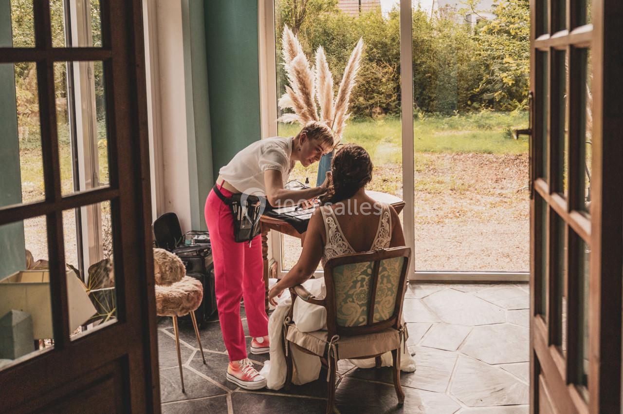 Une maquilleuse applique du maquillage sur une femme assise près d'une fenêtre donnant sur un jardin.