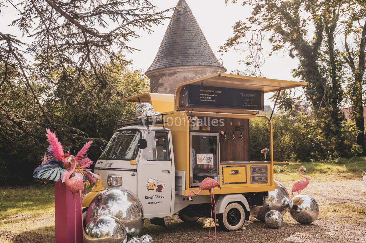 Camion de street food jaune stationné dans un jardin, entouré de flamants roses décoratifs et de sphères argentées.