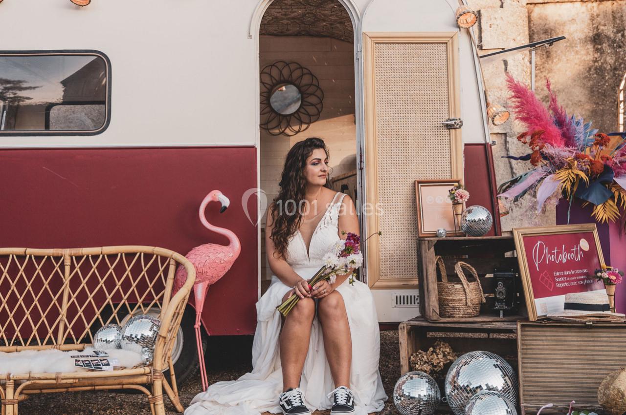 Une femme en robe blanche assise devant une caravane décorée avec des fleurs, des miroirs et des objets rétro.