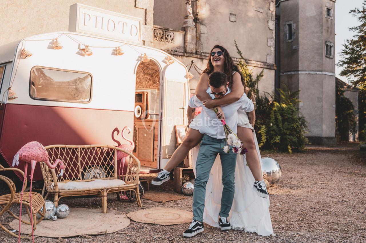 Une femme en robe blanche porte un homme sur son dos devant une caravane rétro décorée, en extérieur.