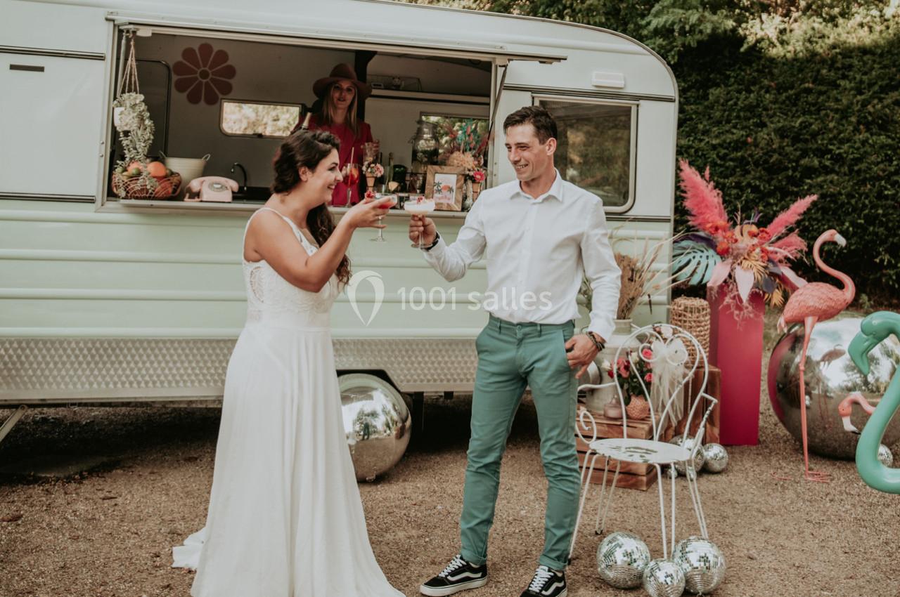 Un couple trinque devant une caravane rétro aménagée, entourée de décorations colorées et festives.