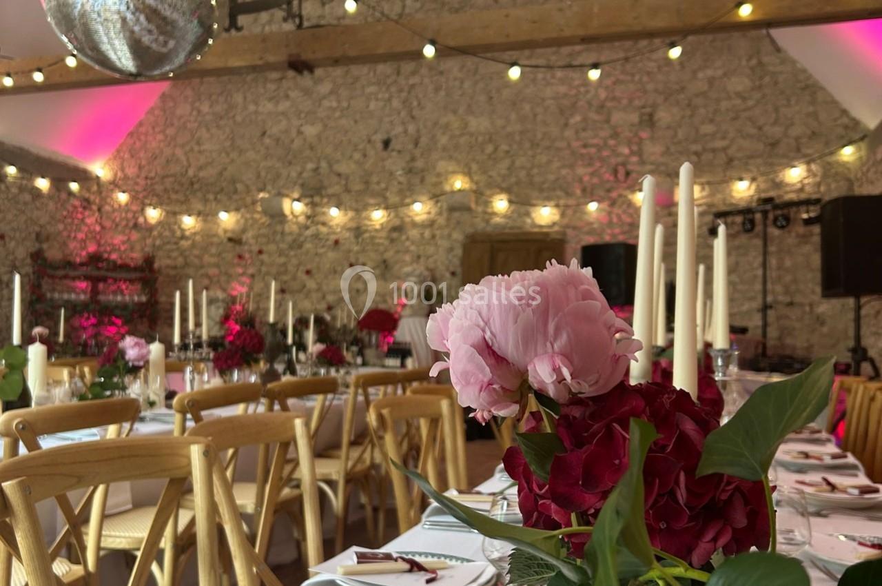 Salle de réception décorée avec des tables dressées, des fleurs roses et rouges, et des guirlandes lumineuses suspendues.