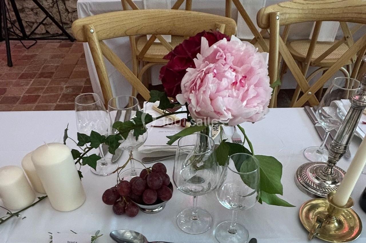 Centre de table avec pivoines roses, bougies, raisins rouges et verres sur une nappe blanche.