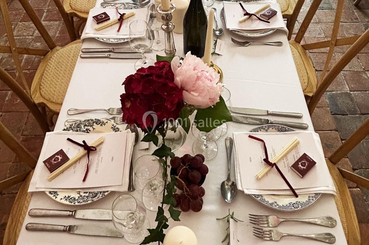Table dressée avec nappes blanches, vaisselle élégante, fleurs, bougies et grappes de raisin sur un sol en pierre.