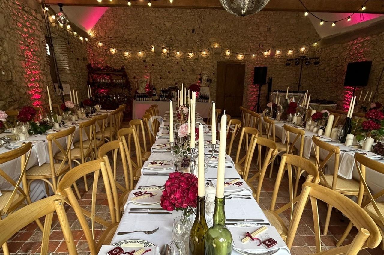 Salle de réception en pierre décorée pour un mariage, avec longues tables dressées, bougies et fleurs rouges.