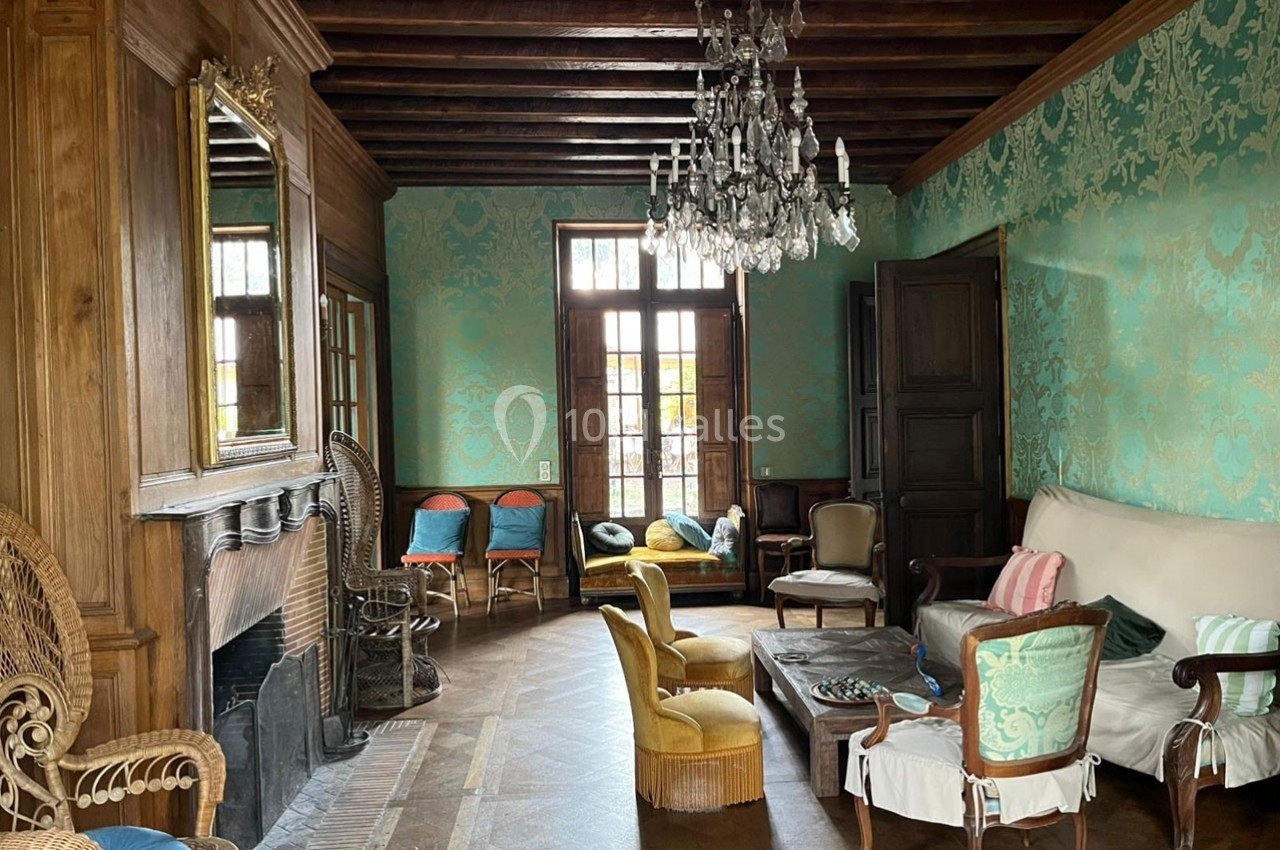 Salon ancien avec murs tapissés, cheminée, mobilier classique et lustre en cristal, éclairé par des fenêtres.