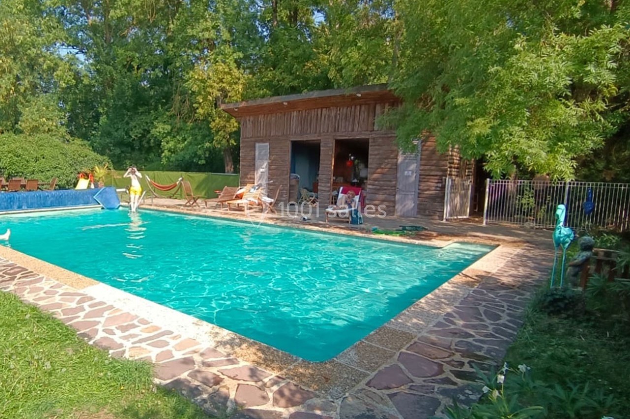 Piscine extérieure entourée de verdure, avec un bâtiment en bois, des chaises longues et des décorations.
