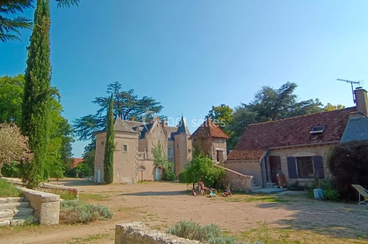 Cour d'un domaine rural avec un manoir en pierre, des dépendances, des arbres et un ciel dégagé en arrière-plan.