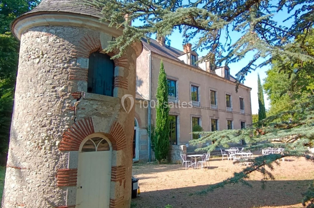 Petit bâtiment en pierre avec une tour ronde, devant une grande maison ancienne entourée d'arbres et de mobilier de jardin.