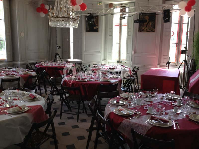 Salle décorée pour un événement avec des tables rondes dressées, nappes rouges et blanches, et un lustre central.