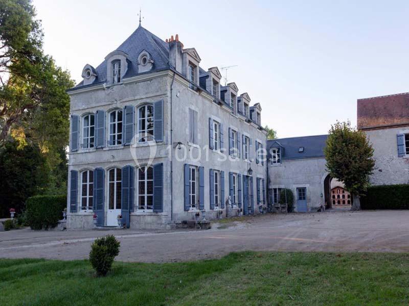 Façade d'un manoir ancien à deux étages avec des volets bleus, entouré d'arbres et d'une cour en gravier.