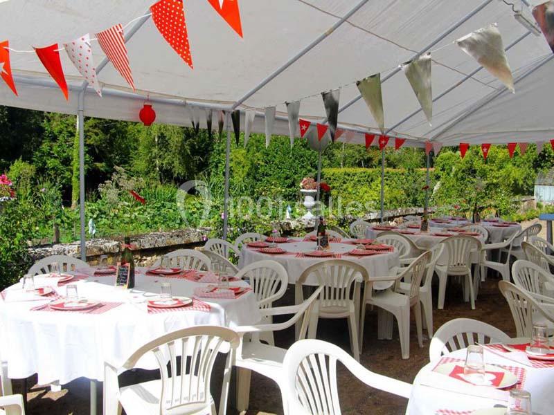 Tables rondes décorées avec nappes blanches et chaises en plastique sous une tente, entourées de verdure.