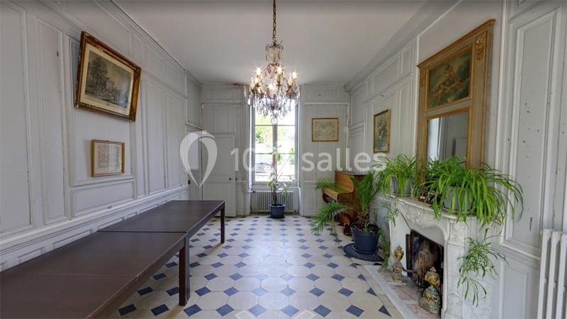 Salle lumineuse avec murs blancs, cheminée décorée, plantes vertes, lustre et table en bois sur sol à motifs.