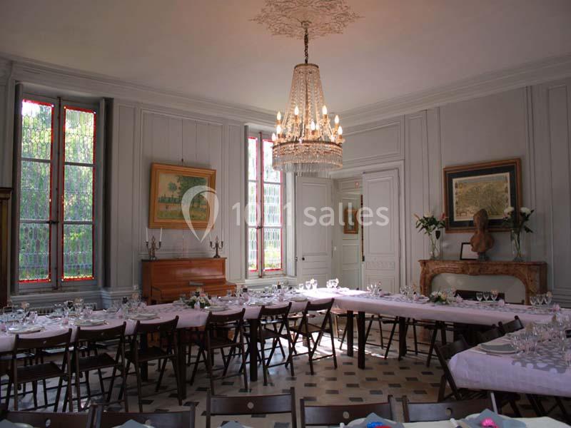 Salle à manger élégante avec de longues tables dressées, un lustre central et des fenêtres laissant entrer la lumière…