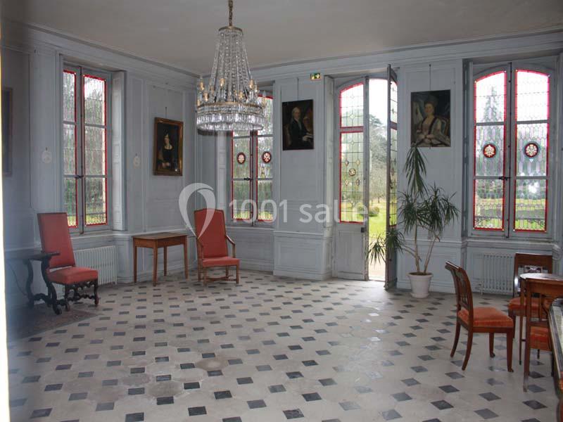 Salle lumineuse avec carrelage en damier, chaises anciennes, lustre en cristal et grandes fenêtres donnant sur un jardin.
