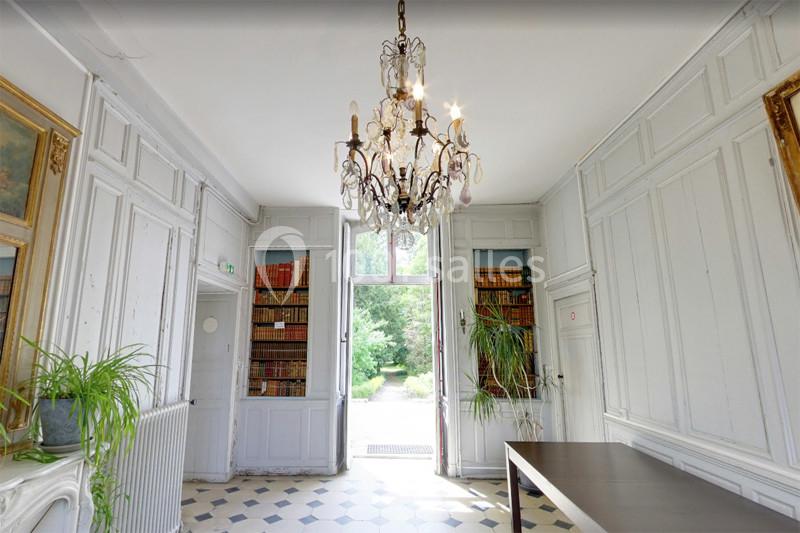 Salle lumineuse avec parquet en damier, bibliothèque intégrée, plantes vertes et lustre central, donnant sur un jardin.