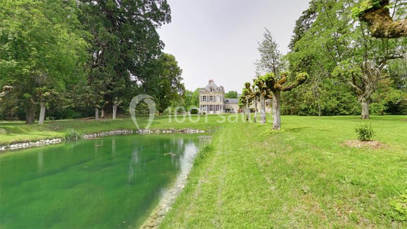 Maison entourée d'un grand jardin verdoyant avec un étang au premier plan et des arbres taillés sur la droite.