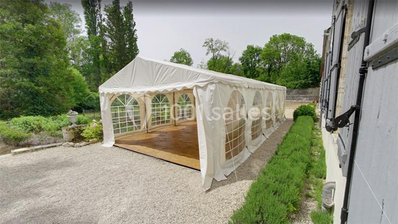 Tente blanche avec sol en bois installée sur une allée gravillonnée, entourée de verdure et d'un bâtiment en pierre.