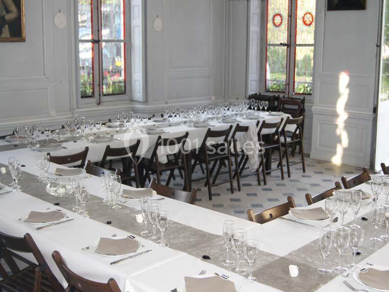 Salle de réception lumineuse avec de longues tables dressées, nappes blanches, verres et serviettes soigneusement disposés.